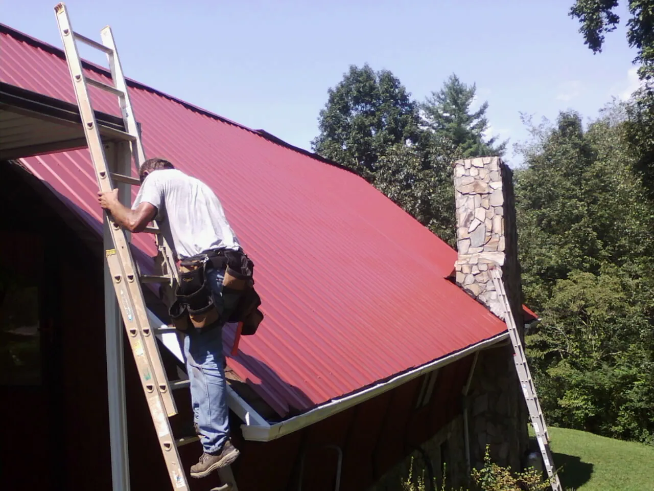 Roofer on a ladder beside a red metal roof and stone chimney — Soffit & Fascia Repair by Roofing Brothers in Bonifay